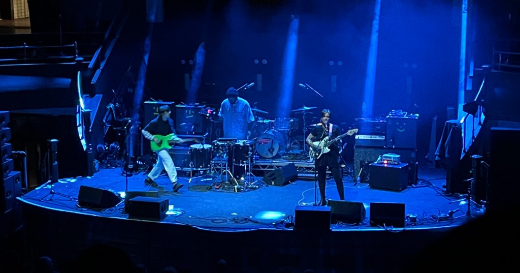 3 members of Dehd performing live on the Albert Hall stage bathed in blue light